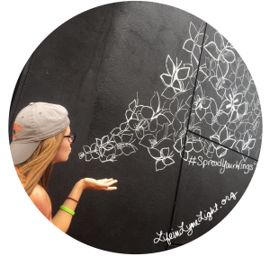 Woman (Lauren, project creator) with backwards cap on and glasses on, blowing kisses with white chalk butterflies drawn on black wall behind her, so it looks like she is creating them. 